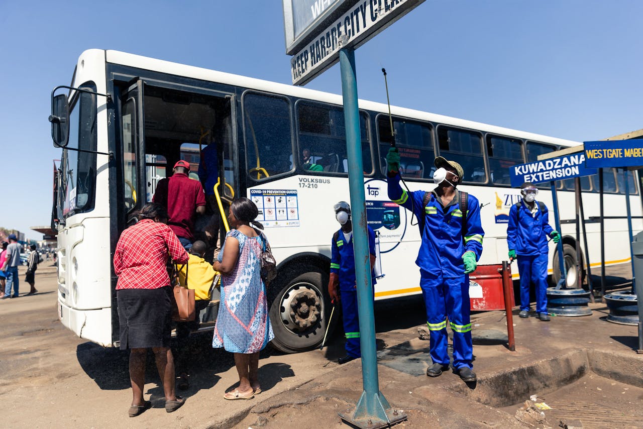 Bij een een bushalte in Harare, Zimbabwe, wordt de omgeving gedesinfecteert met ontsmettende vloeistof. De passagiers proberen onderwijl afstand van elkaar te houden bij de ingang van de bus.